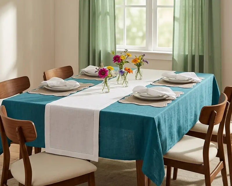 Dining table set with a teal  blue tablecloth, white napkins, and floral centerpieces in a bright room.