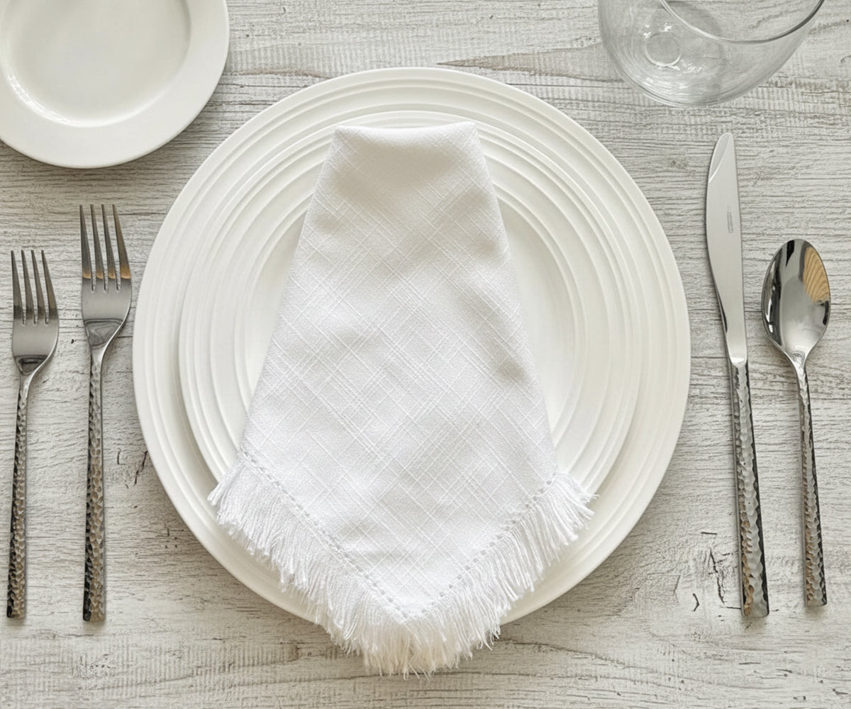 White plates with a folded white napkin, silverware, and glasses on a light wooden table.
