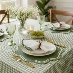 A green and white checkered tablecloth with matching scalloped placemats, white plates, gold-toned cutlery, and a green glass goblet, set in a bright dining room with a floral centerpiece and wooden chairs.