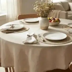 A round dining table covered with a light beige tablecloth, set with white plates, gold placemats, silver cutlery, a folded napkin, and a ceramic vase of dried flowers. The background shows a softly lit living room with a sofa and a bowl of fruit.