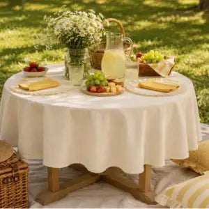 A round table covered with a white tablecloth, set outdoors on grass with plates, a pitcher of lemonade, fruit, and flowers. A wicker basket and striped blanket are partially visible.
