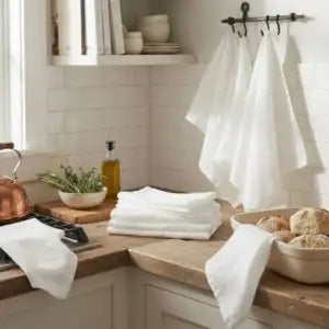 A stack of folded white towels and several hanging white towels displayed on a wall rack in a bright kitchen setting with wooden countertops and white subway tile.