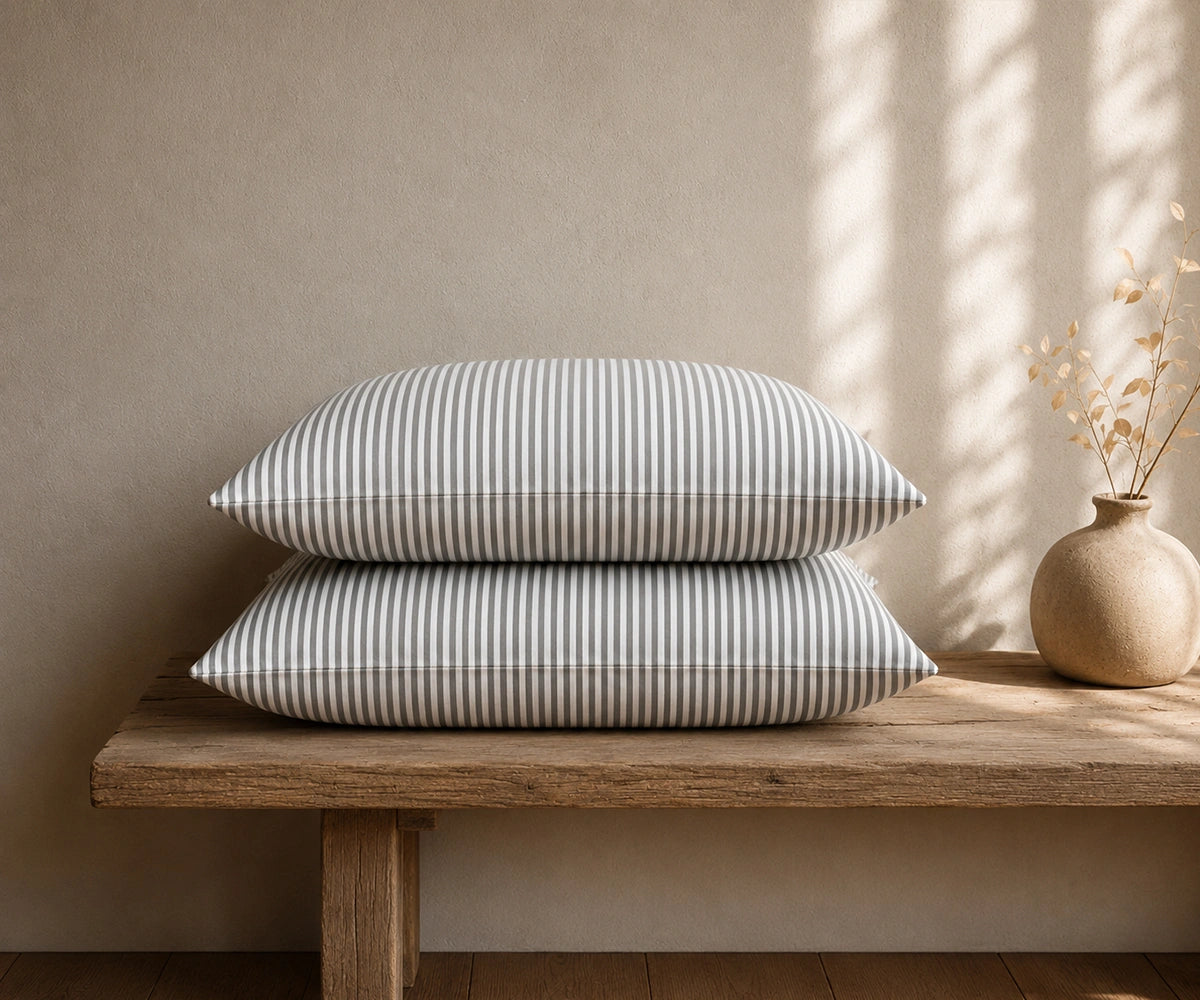Grey and white striped linen pillowcases stacked on a rustic wooden bench with a ceramic vase in a minimal bedroom.