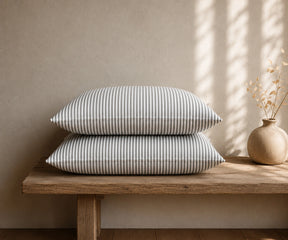 Grey and white striped linen pillowcases stacked on a rustic wooden bench with a ceramic vase in a minimal bedroom.