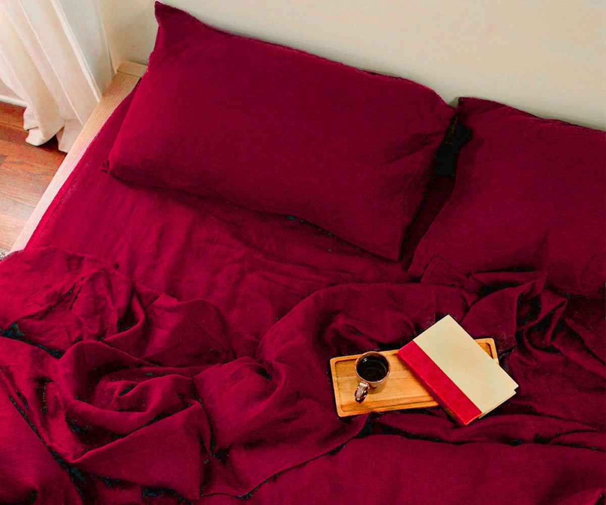 Close-up lifestyle shot of a burgundy linen bedding set featuring textured linen sheets, pillowcases, and a wooden breakfast tray with a coffee cup and book.