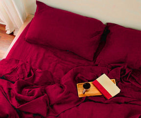 Close-up lifestyle shot of a burgundy linen bedding set featuring textured linen sheets, pillowcases, and a wooden breakfast tray with a coffee cup and book.