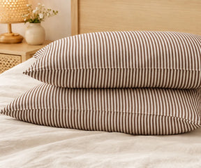 Two stacked chocolate brown and white striped linen pillowcases on a made bed in a bedroom setup