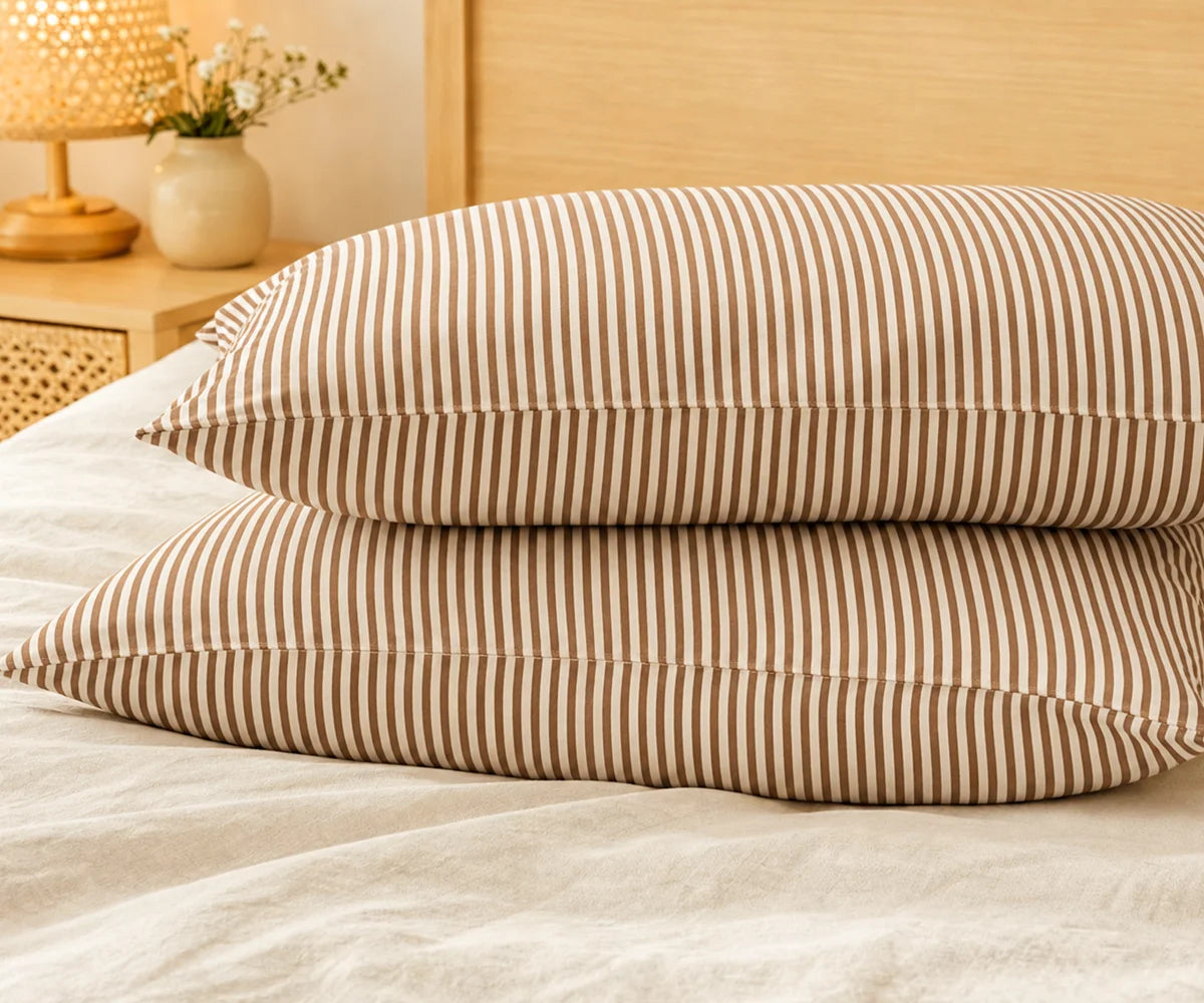 Pair of stacked pillows in coffee brown and white striped linen pillowcases on a beige bed, with a wooden lamp and flower vase on the nightstand.