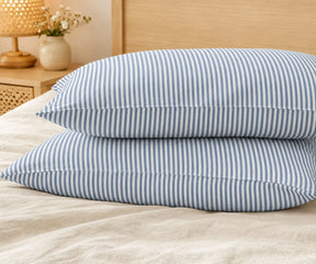 Pair of stacked pillows in dusty blue and white striped linen pillowcases on a beige bed, with a wooden lamp and flower vase on the nightstand.