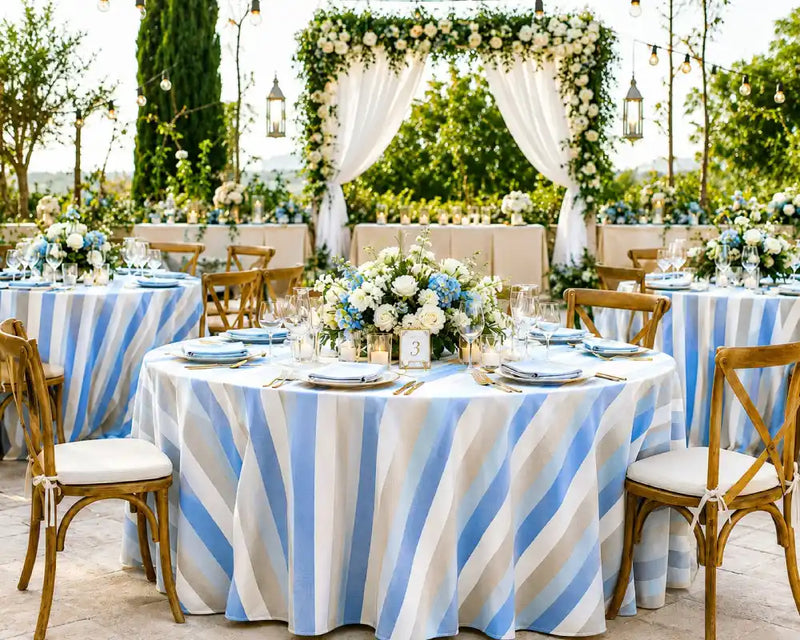 Outdoor event setup with round tables covered in white tablecloths with vertical light blue stripes, set with floral centerpieces, plates, and wooden chairs. A decorated floral arch with white drapery stands in the background among green trees.