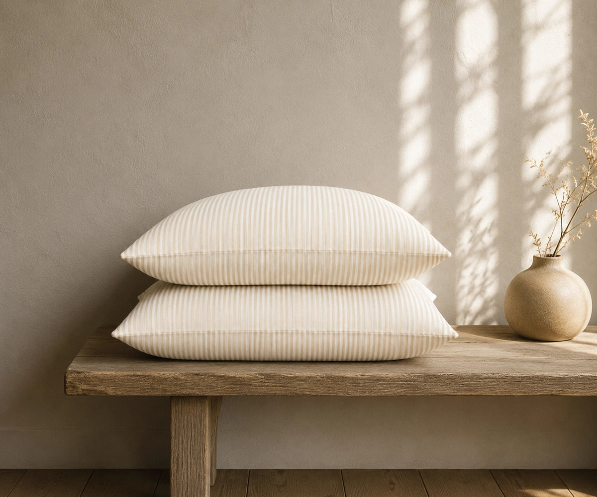 Natural white striped linen pillowcases stacked on a rustic wooden bench with a ceramic vase in a warm, minimal bedroom.