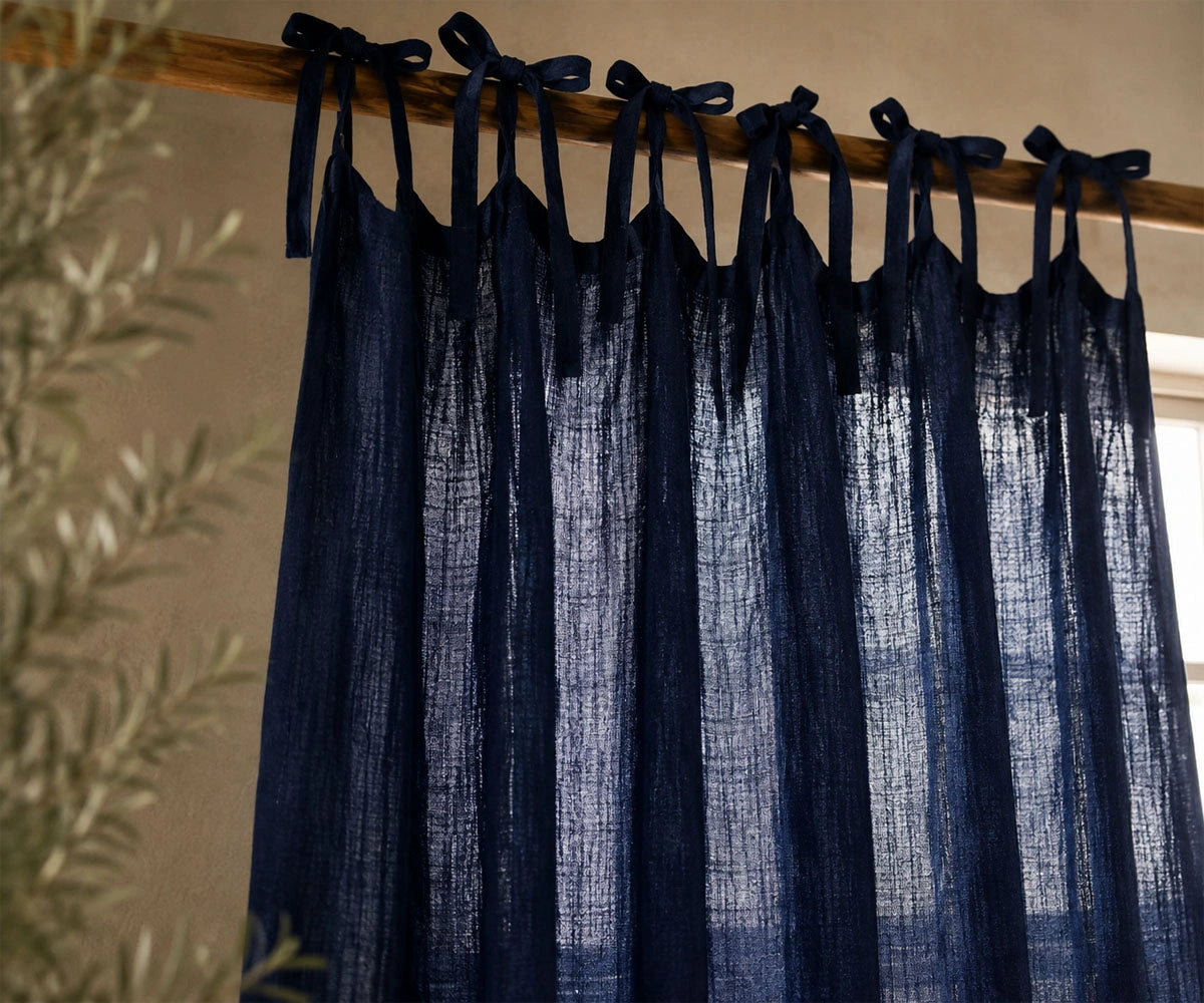 Dark navy blue sheer curtain panels hanging from a wooden rod with matching tie-back loops, shown in a styled interior setting with a blurred plant in the foreground.
