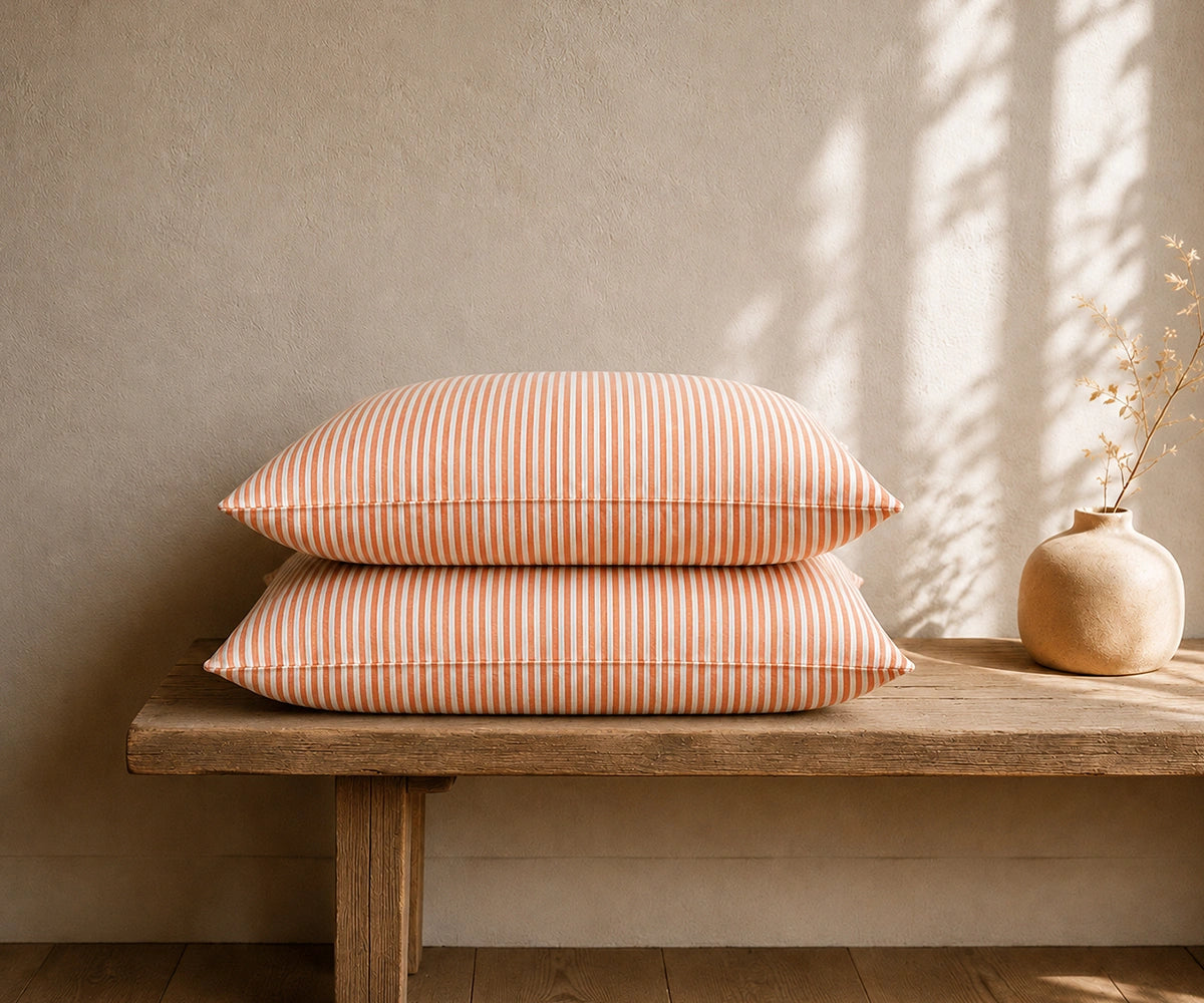 Orange and white striped linen pillowcases stacked on a rustic wooden bench with a ceramic vase in a warm, minimal bedroom.