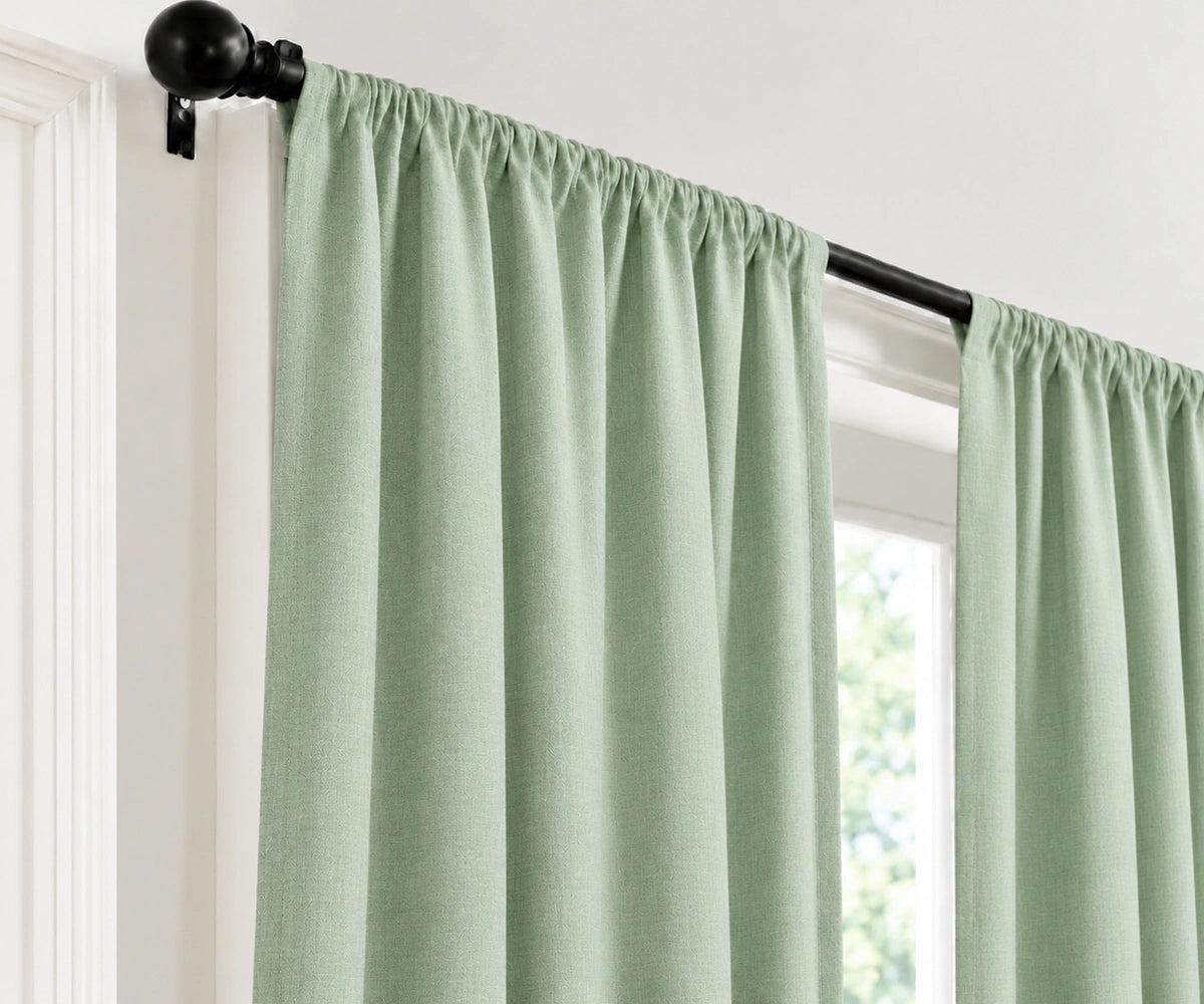 Light sage green curtain panels hanging from a black rod in a window, shown against a white interior wall.