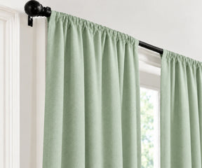 Light sage green curtain panels hanging from a black rod in a window, shown against a white interior wall.