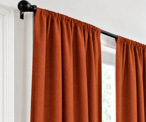 A terracotta curtain panel hanging from a black rod with a black finial, shown in a real interior setting beside a window.