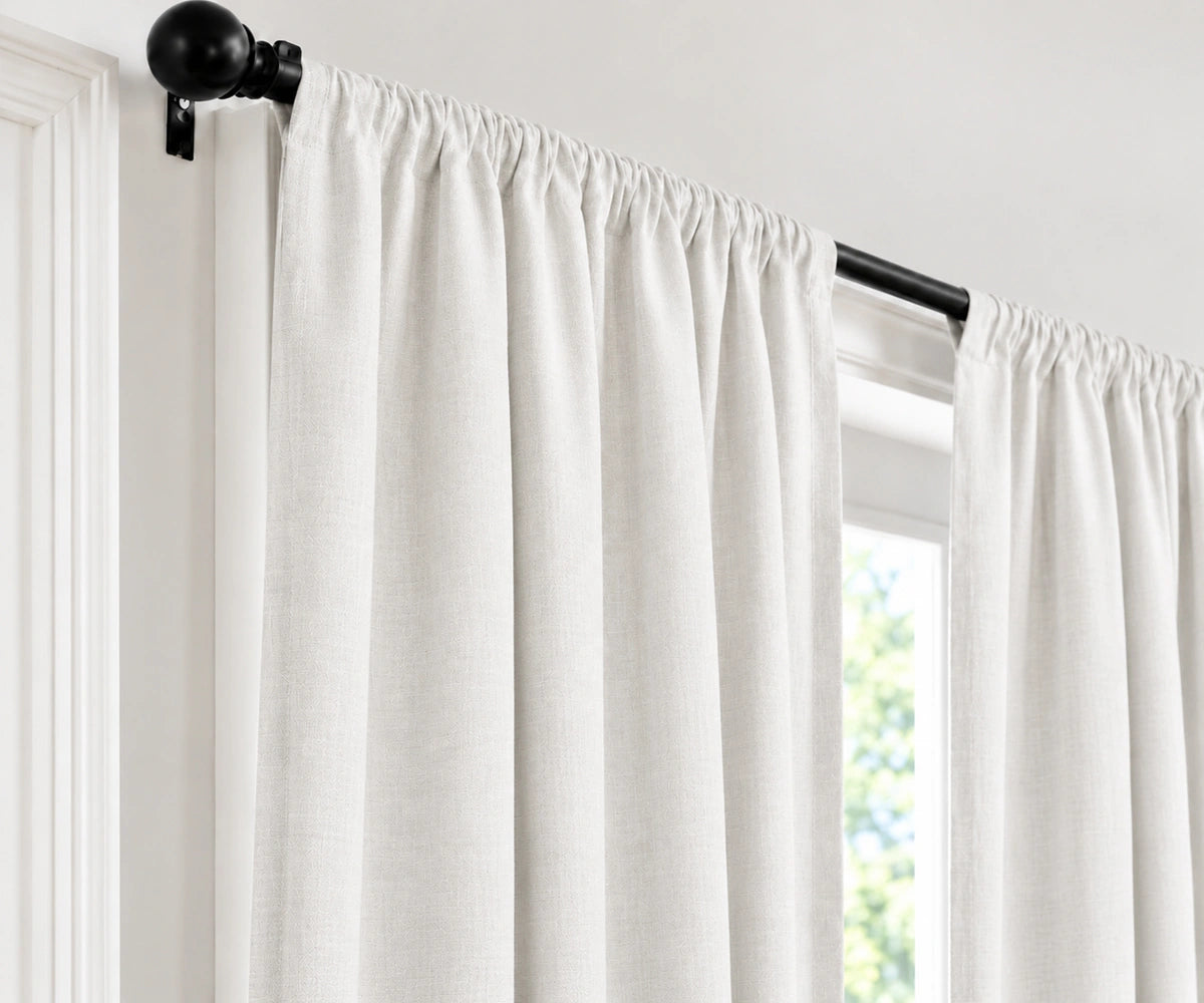 Off-white linen curtain panels hanging from a black metal rod with decorative finials, shown in a bright room next to a window.