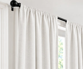 Off-white linen curtain panels hanging from a black metal rod with decorative finials, shown in a bright room next to a window.