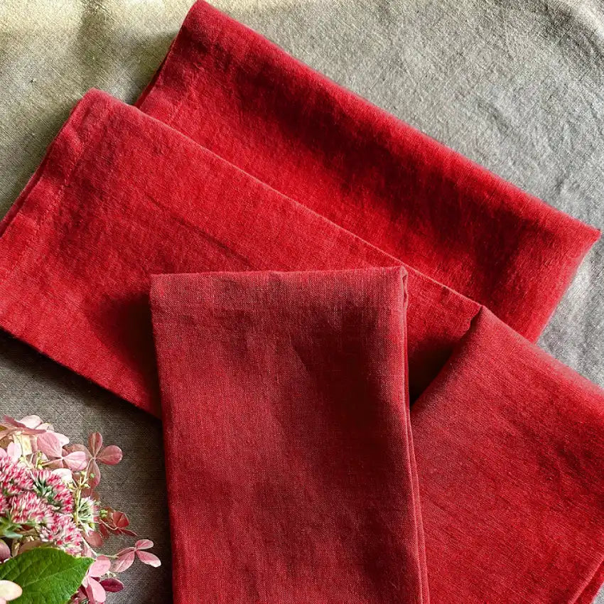 A red linen napkin is placed on a plate, which is set on a wooden table. The napkin has a plain red design and the table setting appears neat and tidy.