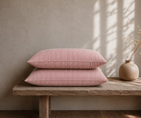  Red and white striped linen pillow covers stacked on a rustic wooden bench with a ceramic vase in a warm, minimal bedroom.