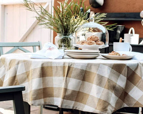 Outdoor table set with a beige and white checkered tablecloth, stacked white plates, pastries under a glass dome, a cup, and a jar with green foliage.