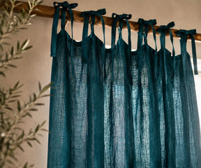 Dark teal blue sheer linen curtains tied to a wooden rod, shown hanging in a room with a beige wall and olive branches on the left.