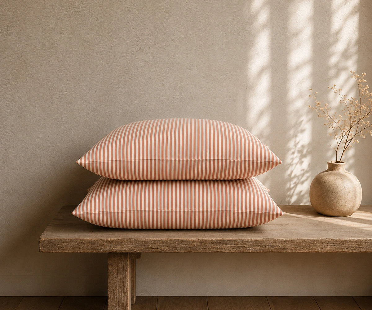 Terracotta and white striped linen pillow covers stacked on a rustic wooden bench with a ceramic vase in a warm, minimal bedroom.