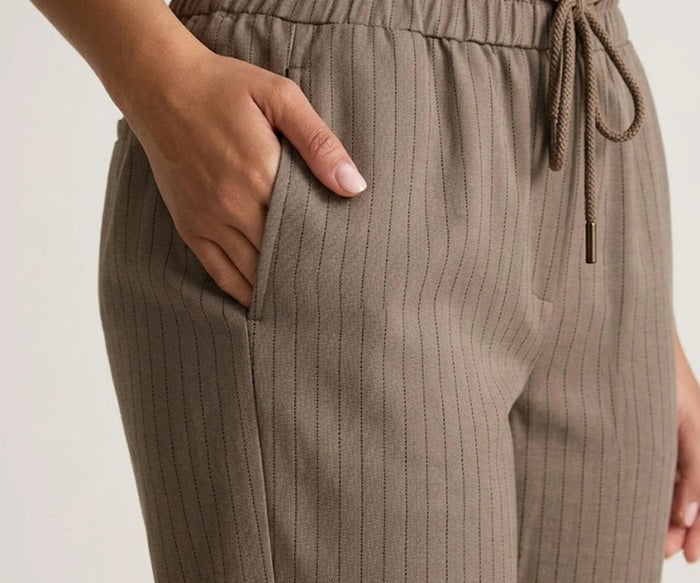 A close-up view of a person sliding their hand into the side slash pocket of dark beige striped linen pants, highlighting the fabric texture and elastic drawstring waist.