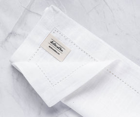 A folded white cotton napkin with borders with the label of All cotton and linen.