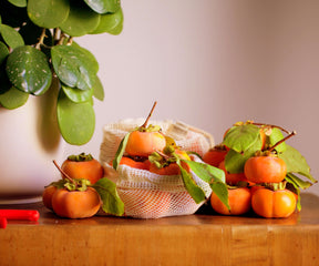 A reusable mesh produce bag with oranges on a kitchen table beside a houseplant